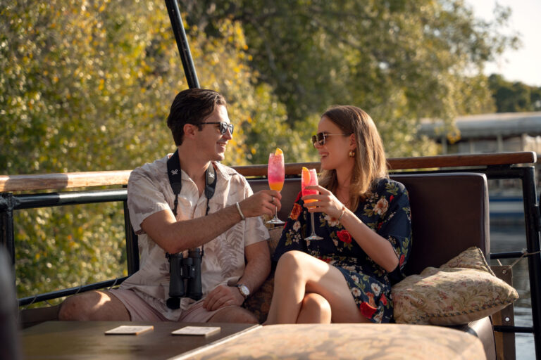 1751964917527_DSC06555 - Go Do Africa Couple toasting colorful cocktails on a boat deck, wearing sunglasses and summer outfits, with trees in the background